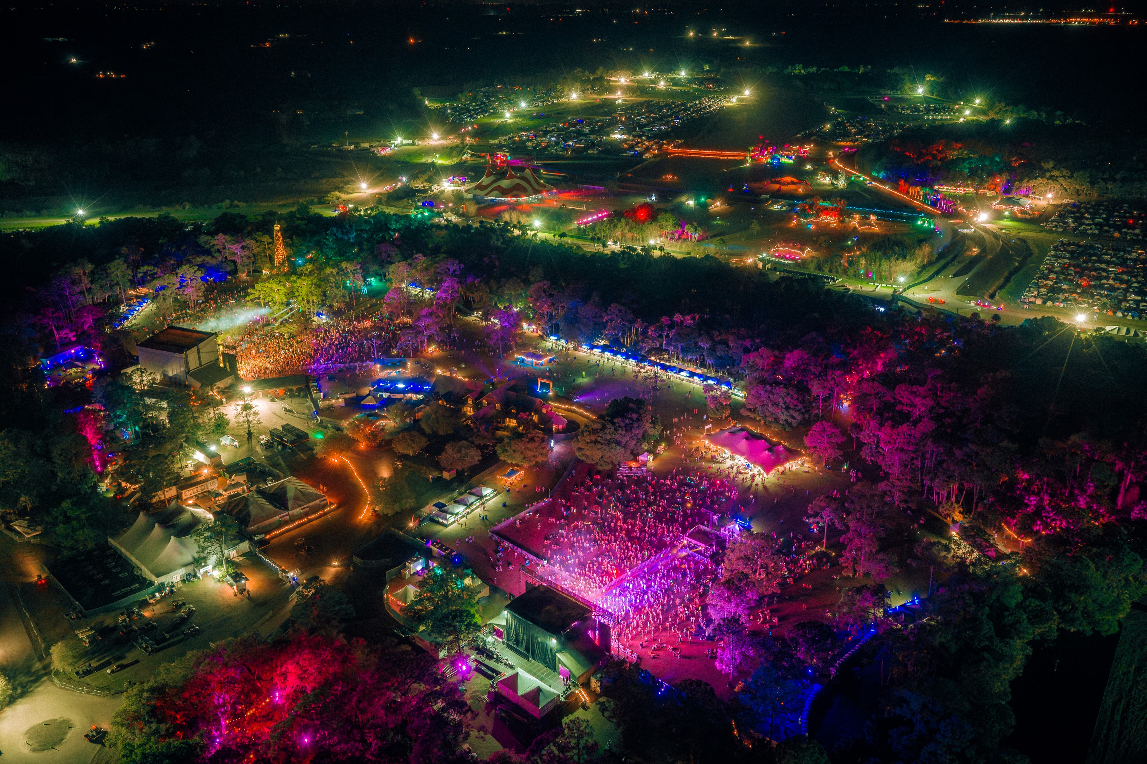 Aerial view of the festival grounds at night, stages and camping lit across the woodland.