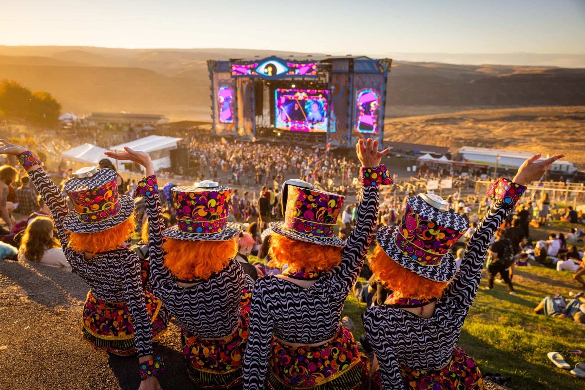 Four attendees in matching costumes raise their arms above a main stage set into a canyon at sunset.